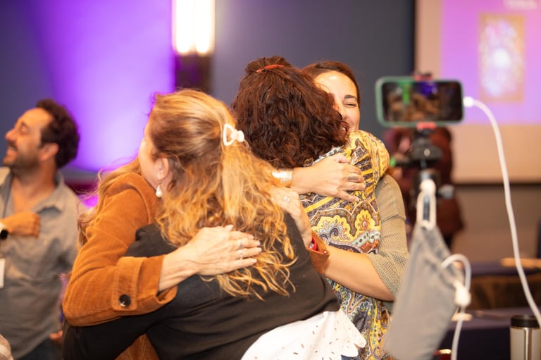 Two women hugging at an indoor event with purple and blue stage lighting, other attendees and camera visible in background