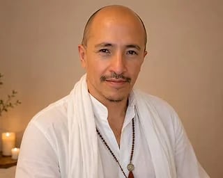Portrait of a bald man with a mustache wearing a white shirt and beaded necklace against a warm neutral background