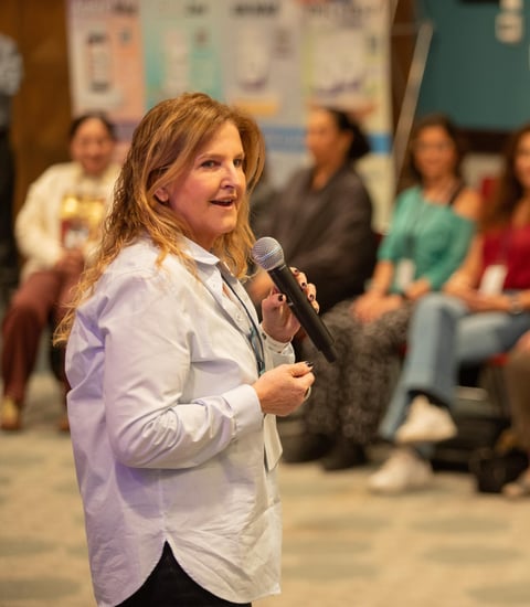 Woman with reddish-blonde hair speaking into a microphone while addressing an audience seated indoors