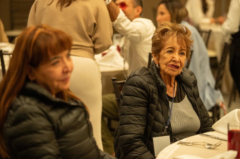 An older woman in a black jacket sits at a table during an indoor event, with other attendees visible in the background