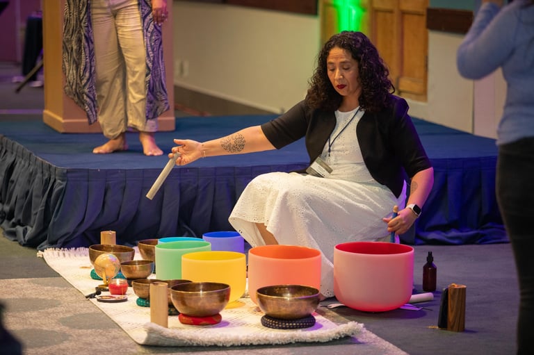 Woman in white dress demonstrating sound bowls and instruments on stage with colorful bowls and singing implements