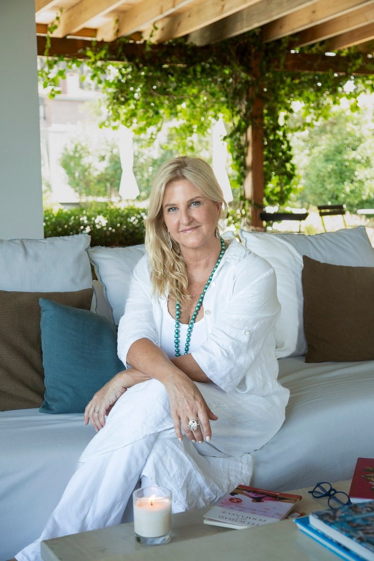 Woman in white linen clothing sitting on a couch in a covered outdoor patio with greenery and beams overhead