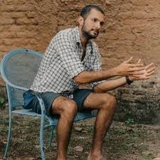 Man in checkered shirt sitting on blue chair stretching arm against brick wall