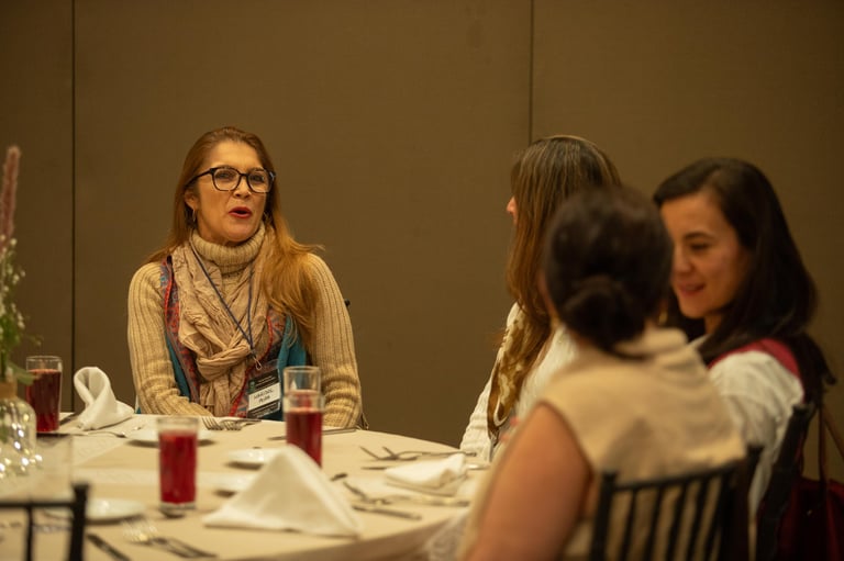Woman wearing glasses and scarf speaking at dinner table with three other women in formal dining setting
