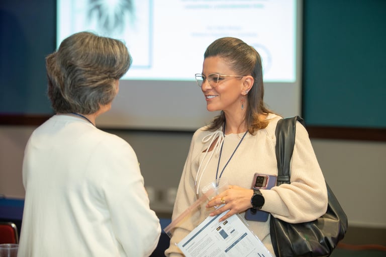 Two women having a conversation at a professional event, one wearing glasses and a credential badge, with a projection screen visible in the background.
