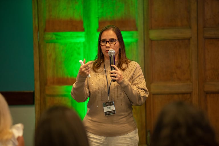 Woman speaking at podium with microphone wearing glasses and name badge, green neon lighting in background