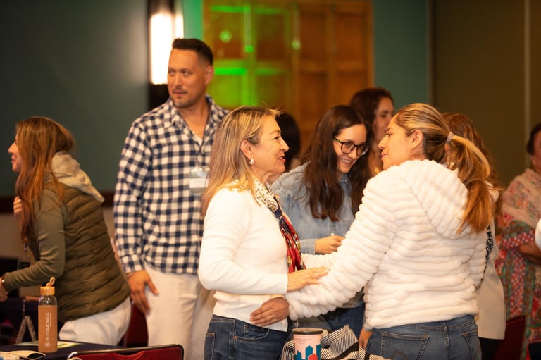 Group of people gathered indoors shaking hands and conversing, appearing to be at a social or community event.