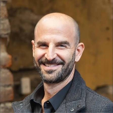 Portrait of a bald man with dark beard wearing a gray jacket and black shirt, smiling at the camera