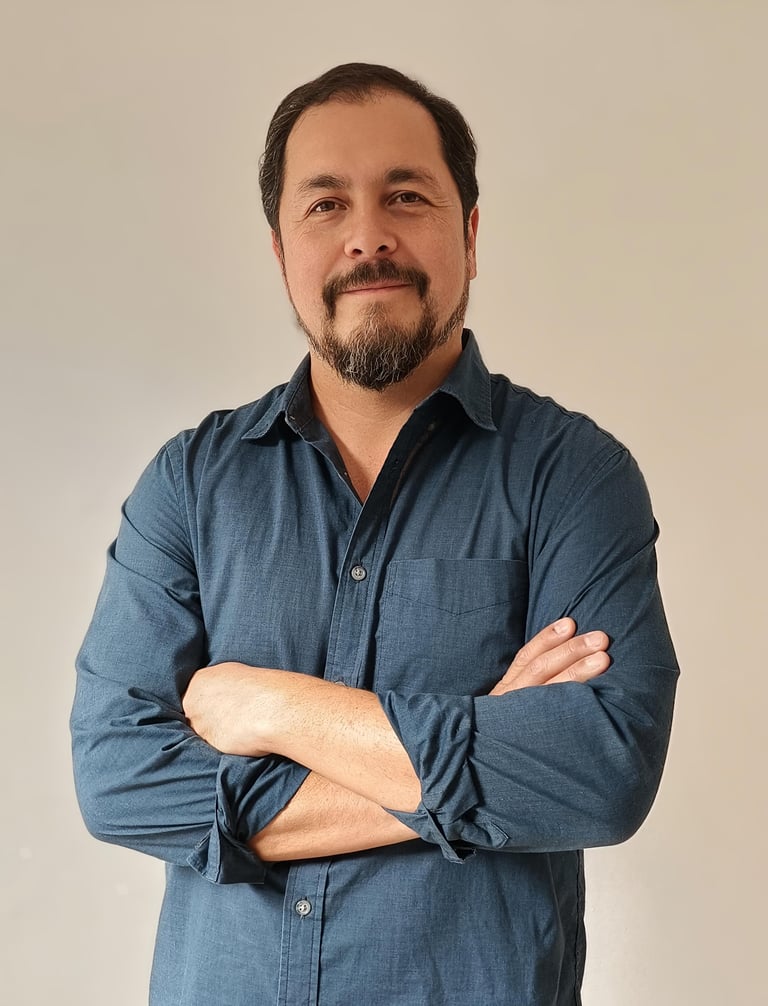 Portrait of a man with dark hair and goatee wearing a blue button-up shirt with arms crossed against a neutral background