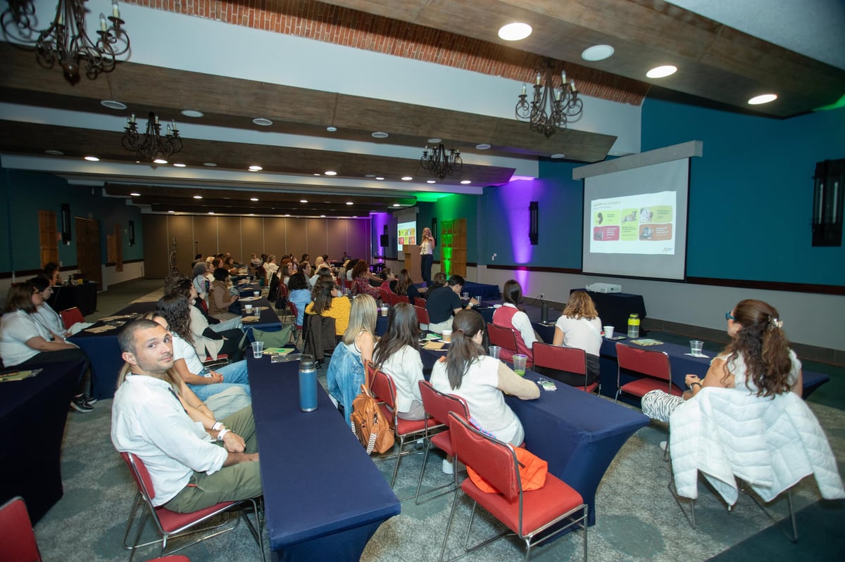 Conference attendees seated at tables in a modern auditorium with colorful stage lighting and projection screen