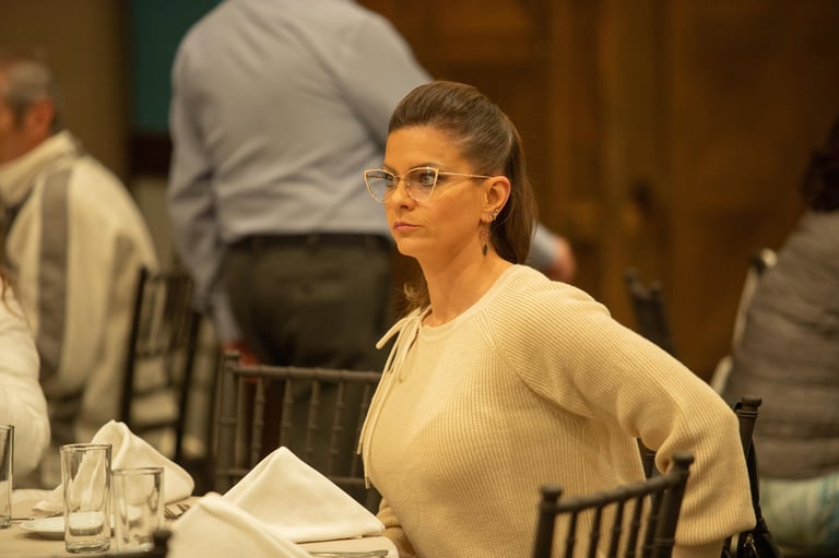 Woman wearing glasses and beige sweater seated at a formal dining table with other people in the background