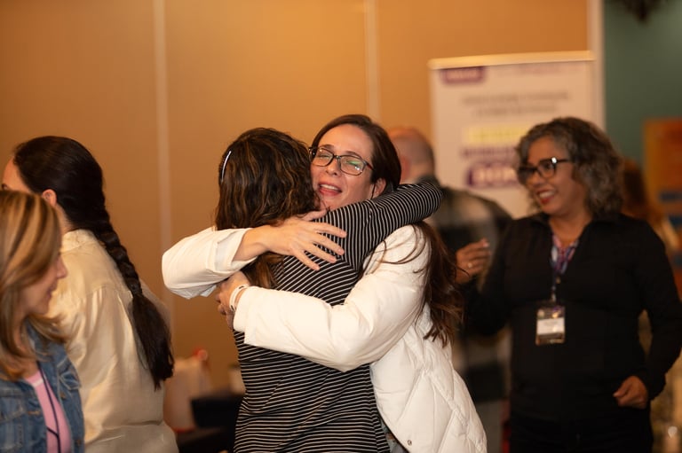 Two women hugging at a professional conference or networking event, surrounded by other attendees in an indoor venue